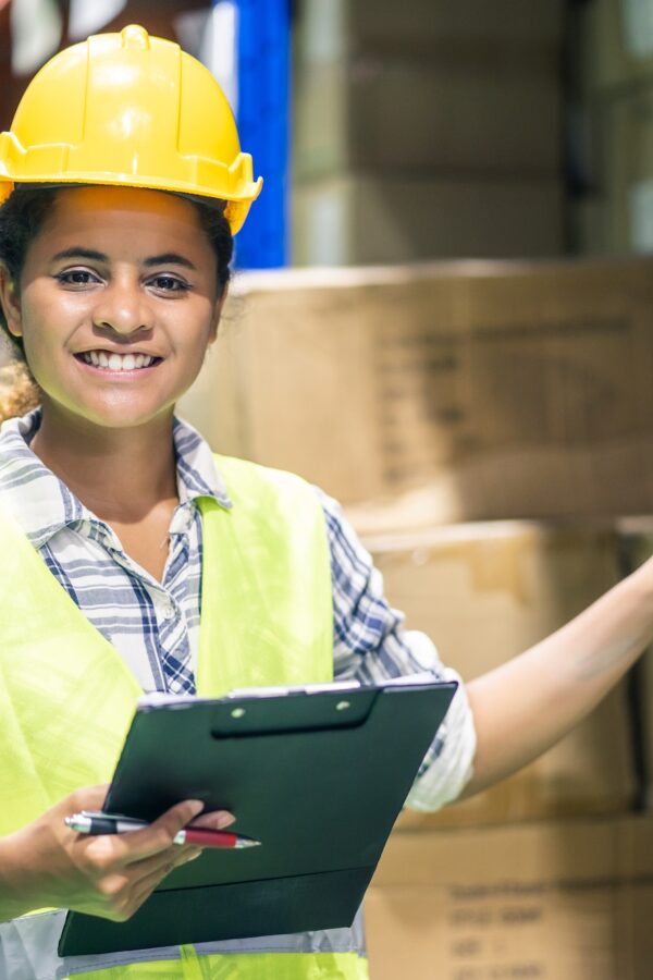 Young woman worker in safety helmet and vest checking box in stock in warehouse.